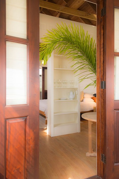 mahogany doors glow softly as they frame the view past an arched palm frond to the open shelving column that separates the keeping room from the bedroom of the Dream Suite at Mahogany Bay Village on Ambergris Caye in Belize