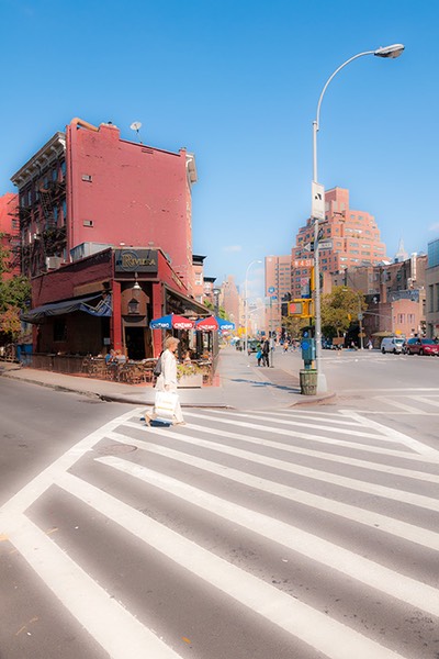 white-clad woman strides across striped crossing, set against backdrop of quirky flatiron building and Manhattan avenue beyond, all guarded by reluctant cobra-head street light